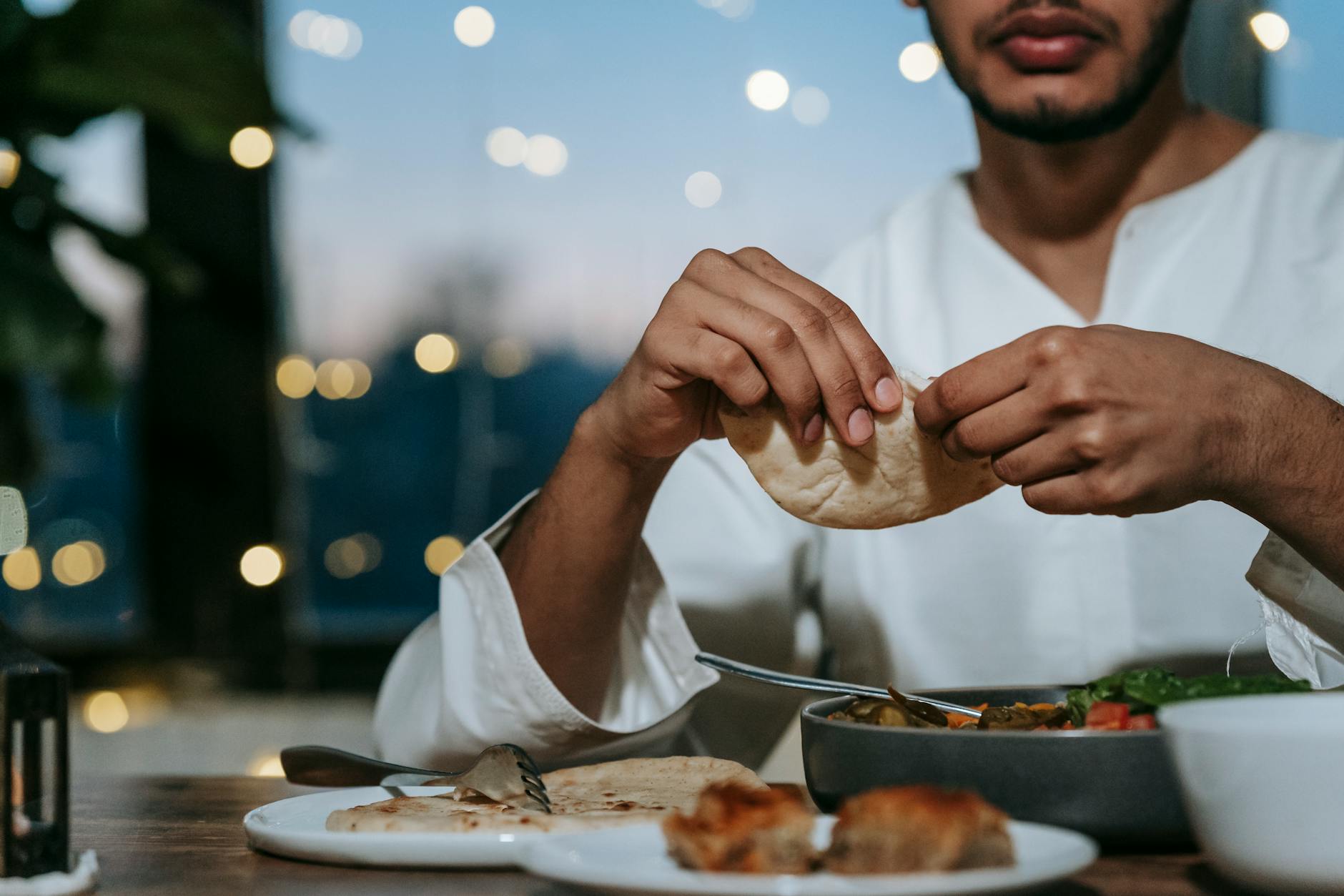 Muslim man breaking pita bread at a dining table for dinner during iftar indoors.