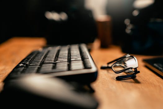 Focus on keyboard and eyeglasses on a warm-lit wooden office desk.