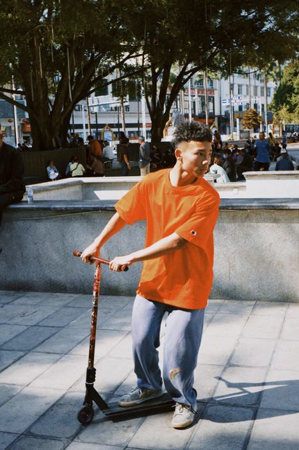 Young male on scooter in urban skate park, wearing bright orange shirt, exploring leisure activities.