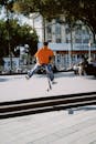 A Man in Orange Shirt and Denim Pants Jumping on the Park while Holding His Scooter Trolley
