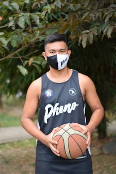 A young man in a basketball jersey and mask holding a ball outdoors.