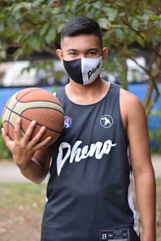 A young man wearing a mask and jersey holding a basketball outdoors, ready to play.