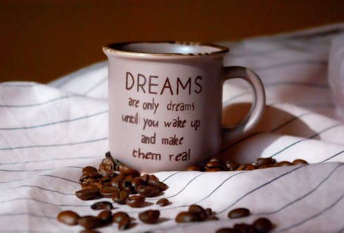 Mug saying Dreams Are Only Dreams Until You Wake Up and Make Them Real on striped fabric with scattered coffee beans on brown background