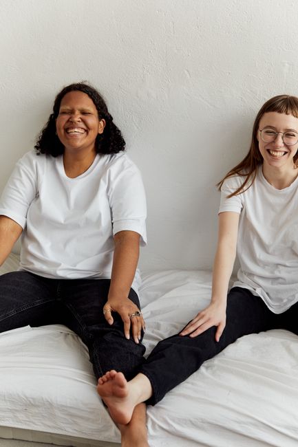Joyful lesbian couple wearing casual attire laughing together indoors on a bed. Bright and warm atmosphere.