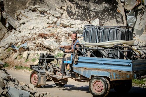 Free Smiling man driving a makeshift vehicle through rubble-strewn street in Idlib, Syria under bright sunlight. Stock Photo