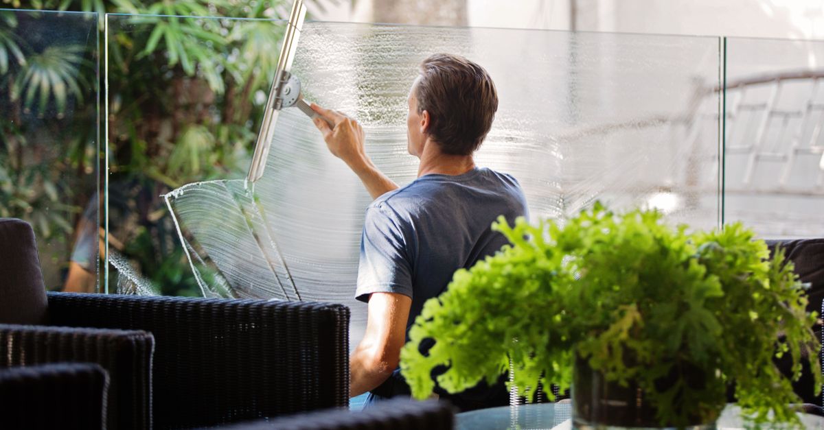 A man cleaning a glass barrier outdoors with a squeegee, surrounded by greenery.