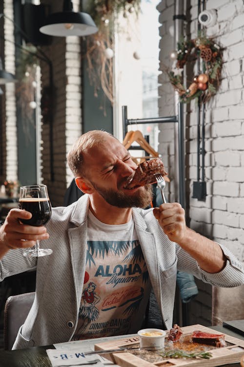 A Man Biting a Steak while Holding a Glass of Beer