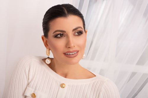 Free Attractive female with black hair and earrings dressed in white elegant clothes looking up while standing near white tulle in room Stock Photo