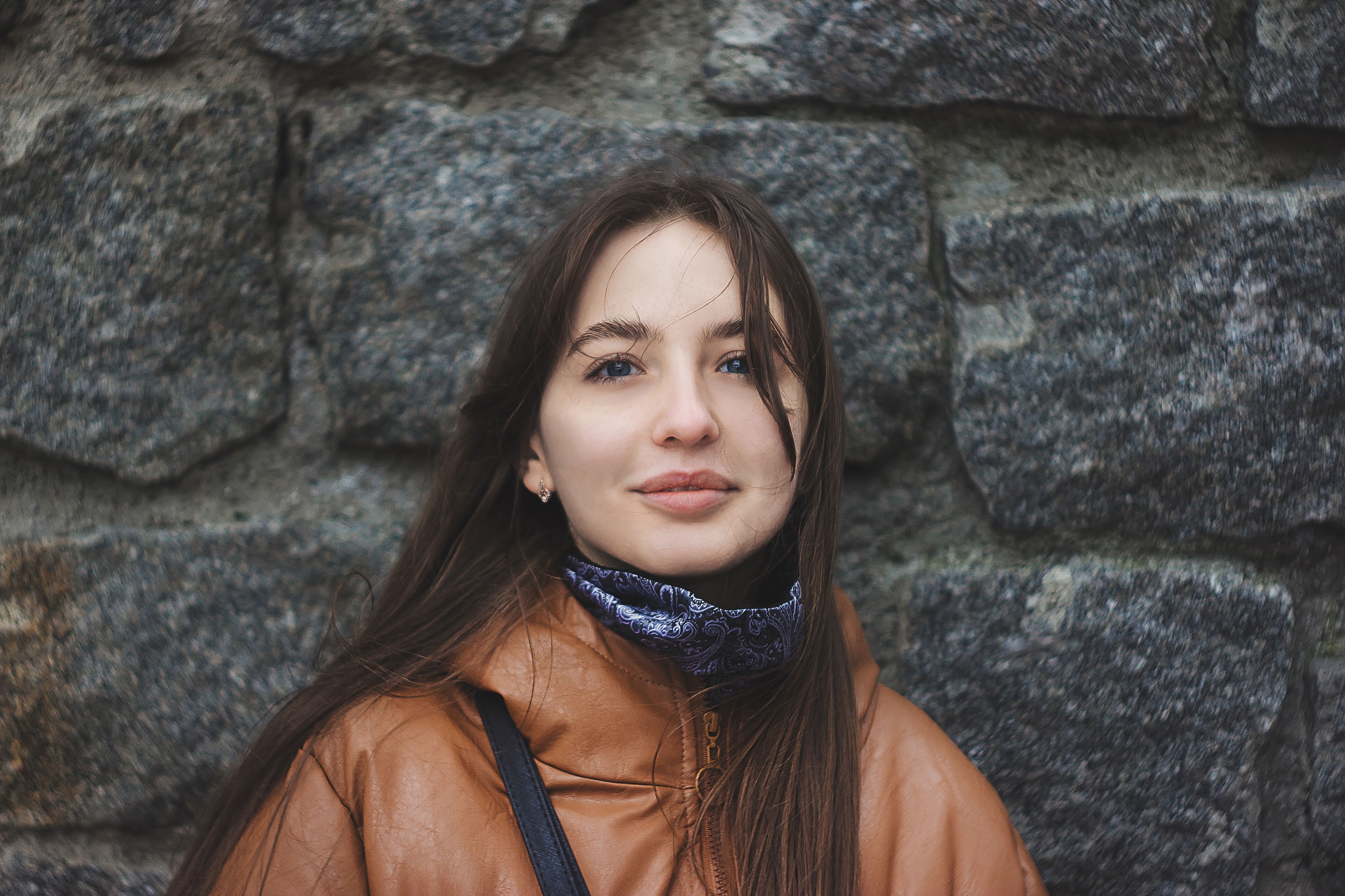 Close-up portrait of a young woman in a leather jacket standing against a stone wall.