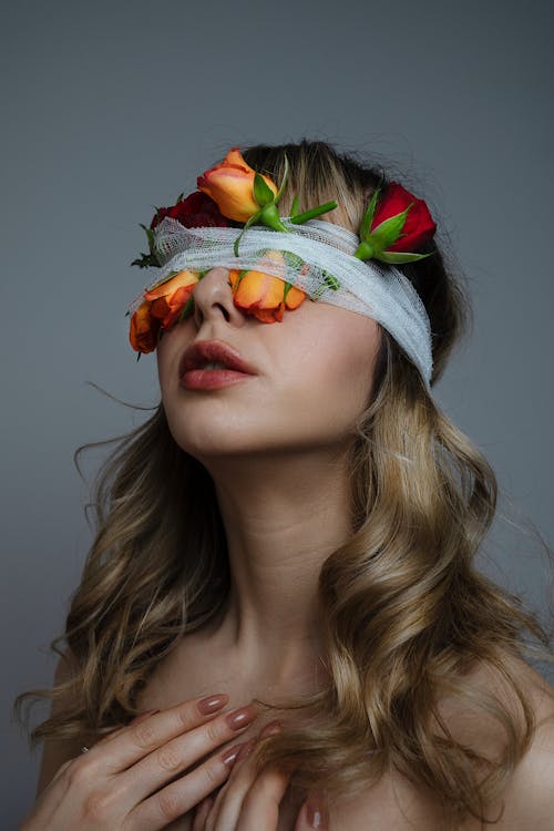 Charming female with long wavy hair and covered eyes with bright blooming flowers standing on gray background in modern studio