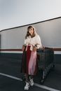 Full body of confident female in stylish outfit looking at camera while leaning on heap of shopping trolleys on parking lot