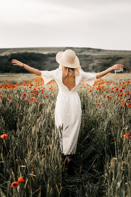 Free Back view full body of stylish female in white dress and headwear strolling with spread arms on grassy meadow with blooming flowers Stock Photo
