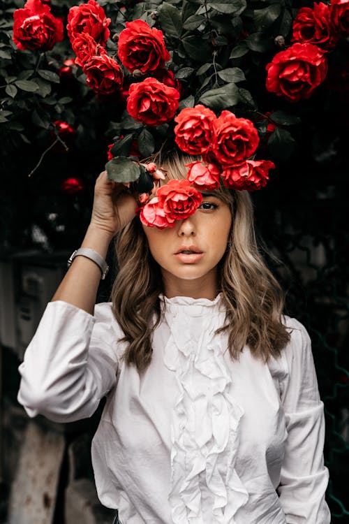 Free Attractive female looking at camera and covering eye with red flowers while standing under branches of blooming bush in garden Stock Photo