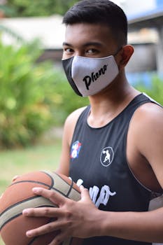 A young man holding a basketball and wearing a face mask in an outdoor setting, ready to play.