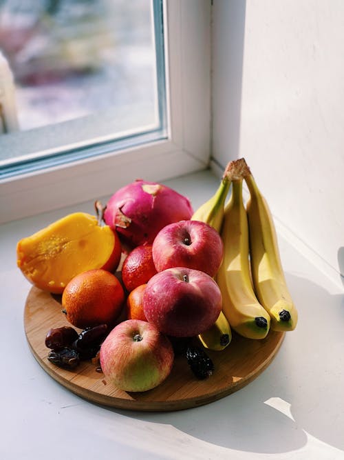 Wooden plate with apples and oranges with bananas together with mango and dragon fruit on white windowsill