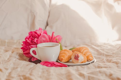 Cup of fresh aromatic coffee with plate of yummy croissants with berry jam placed on soft bed with gentle peony in sunny morning