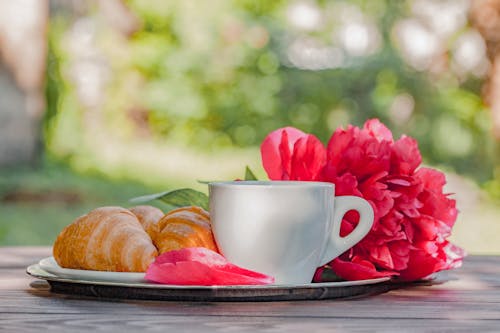 Ceramic cup of hot coffee with plate of crunchy croissants served on tray with fresh fragrant pink peony on wooden table on veranda
