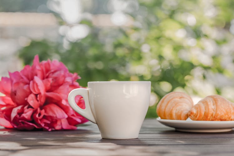 Coffee Cup With Croissants Served On Table Near Peonies In Green Garden In Sunlight