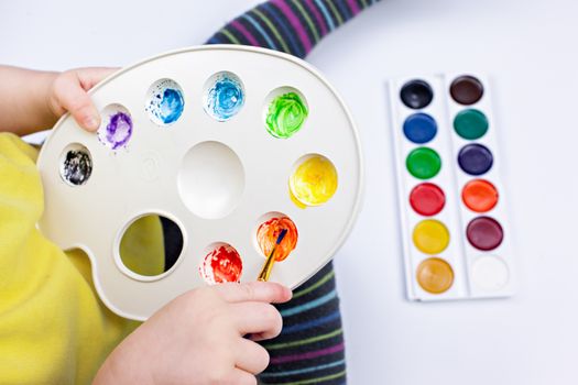From above of crop unrecognizable kid with paintbrush and palette blending aquarelle paints on white background