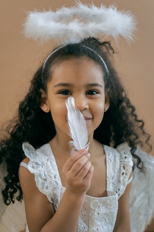 Adorable African American girl with white feather wearing angel costume with halo looking at camera on brown background in studio