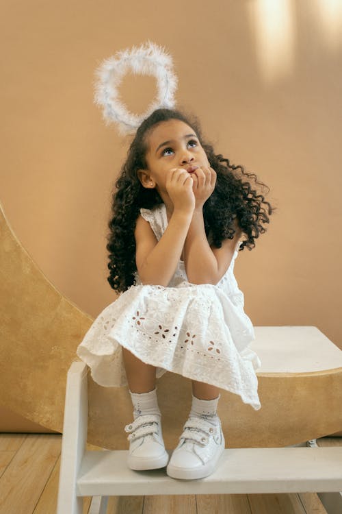 Free Full body of adorable African American girl in white angel costume with halo looking up while sitting on white step on brown background Stock Photo