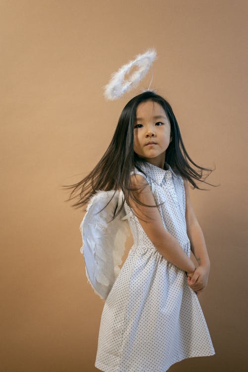 Free Cute Asian girl wearing white angel costume with wings and looking at camera while shaking hair on brown background in studio Stock Photo