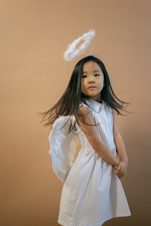 Cute Asian girl wearing white angel costume with wings and looking at camera while shaking hair on brown background in studio