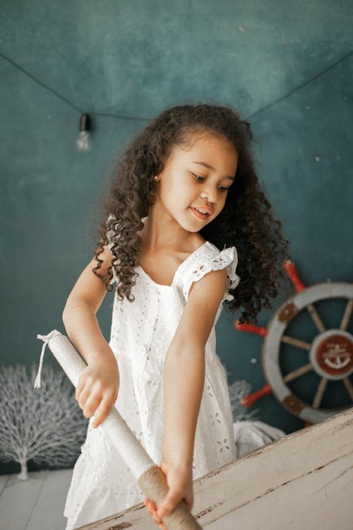 Adorable little African American girl in white dress with wooden paddle playing in studio with nautical decor