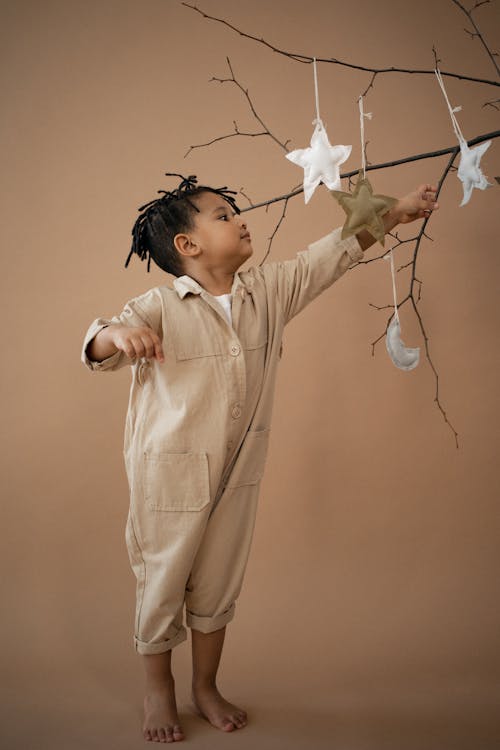 Side view full body of cute barefoot African American boy standing near leafless sprigs with white decorative stars on brown background