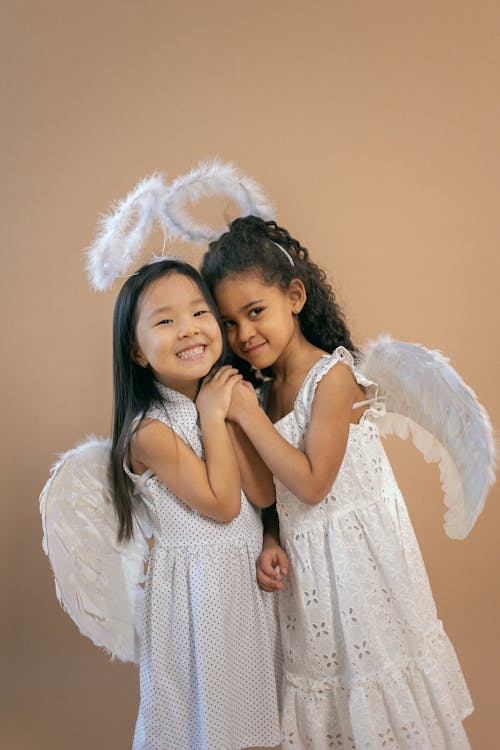 Positive multiracial girls with black hair in white angel costumes with wings and halos standing on brown background in light studio