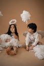 Full body of diverse children in angel costumes with halo and wings sitting on brown background in studio while playing with white cotton wool