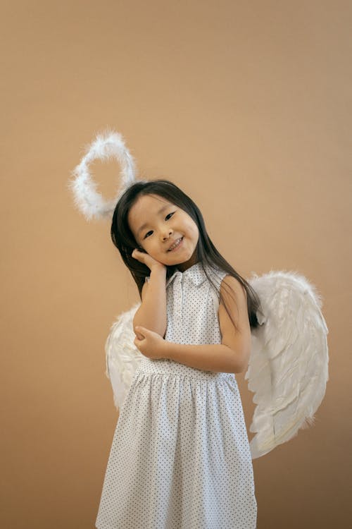 Adorable Asian girl in angel outfit with wings and nimbus leaning on hand while looking at camera on brown background