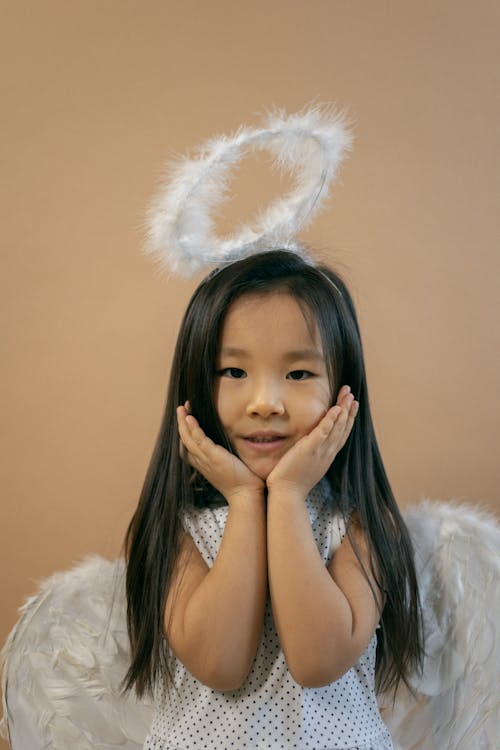 Content Asian girl in white wings and nimbus looking at camera while leaning on hands on brown background in studio