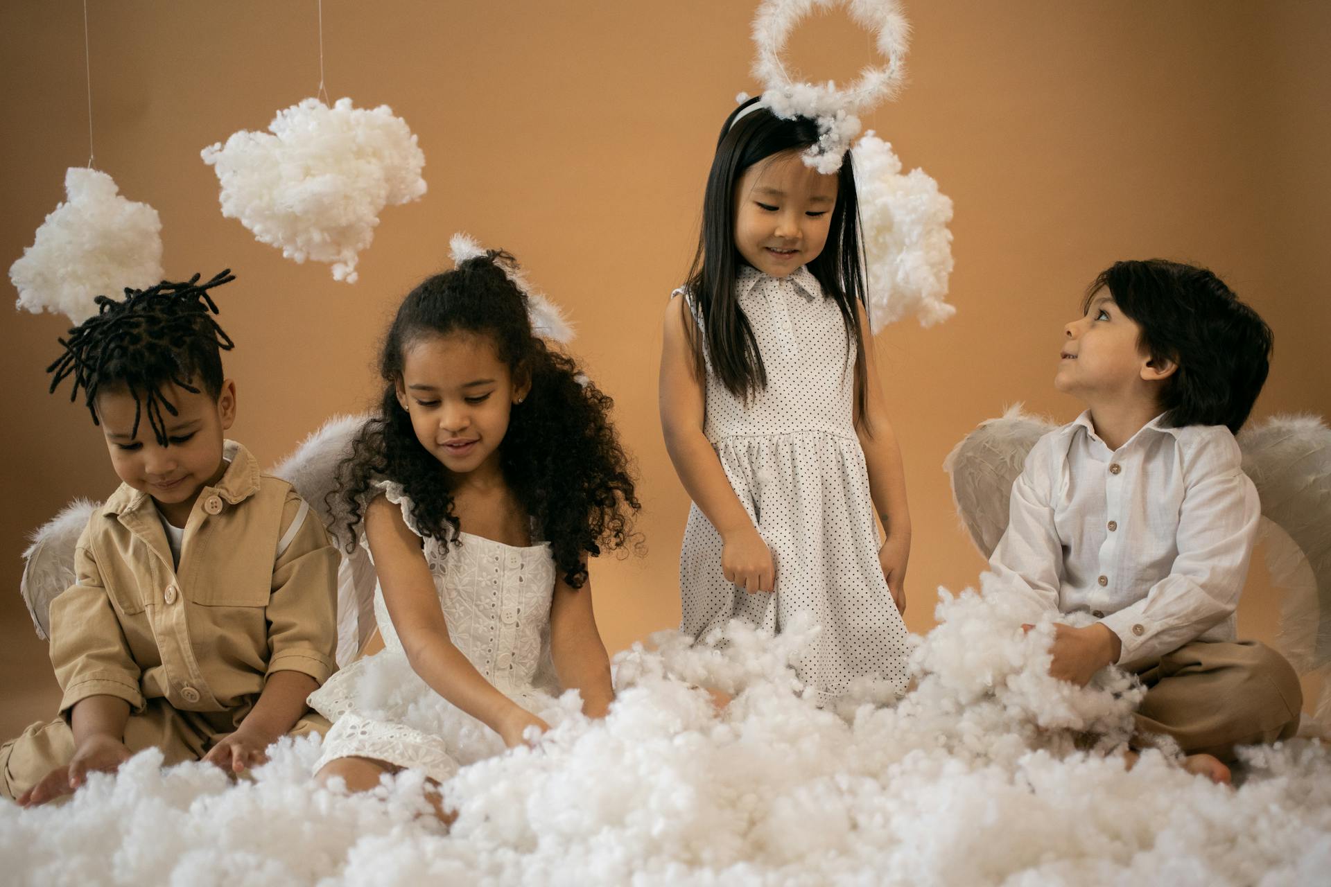 Positive multiracial children in angel outfits with halo and wings playing with decorative with cotton wool while sitting on brown background