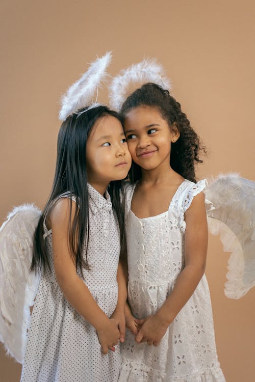 Free Content multiracial girls wearing similar angel costumes with wings and halos looking at each other and touching faces while standing on brown background Stock Photo