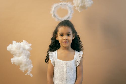 Cute African American girl in white dress and halo looking at camera while standing on brown background with decorative clouds
