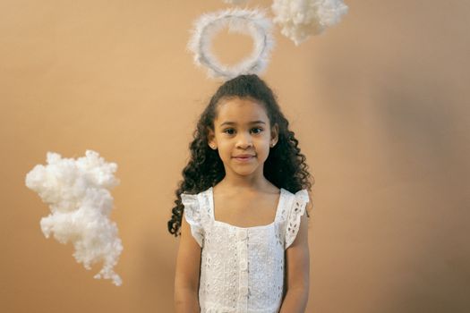 Cute African American girl in white dress and halo looking at camera while standing on brown background with decorative clouds