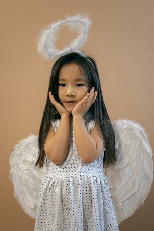 Adorable Asian girl in white angel costume with wings and nimbus looking at camera while leaning on hands on brown background