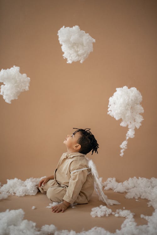 Side view of adorable African American boy in white wings looking up while sitting on brown background with decorative clouds made of cotton wool