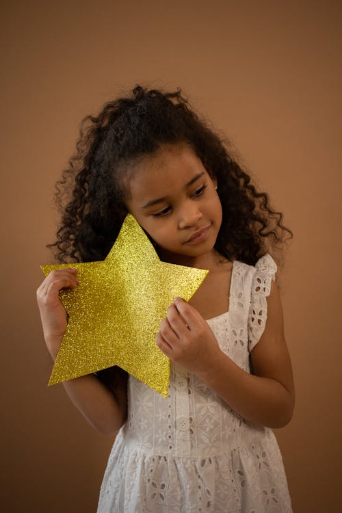 Free Adorable African American girl in stylish white dress with decorative yellow star in hands standing on brown background in studio Stock Photo