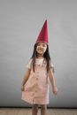 Delighted Asian girl in red gnome cap smiling and looking at camera while standing on gray background in light studio