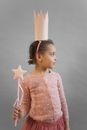 Cute African American girl wearing princess costume with crown and magic wand while standing on gray background in light studio