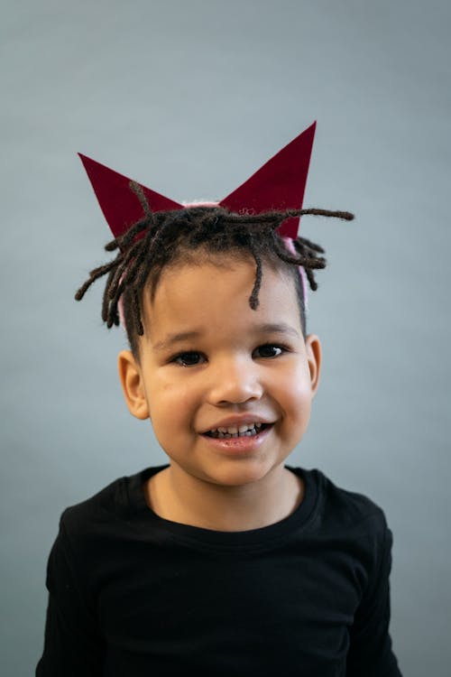 Free Delighted African American boy with red horns smiling and looking at camera while standing on gray background in light studio Stock Photo