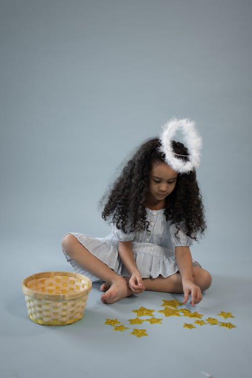 Full body of barefoot African American girl in angel outfit with halo sitting on floor while playing with decorative yellow stars on gray background