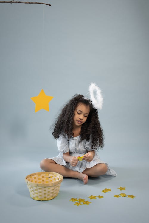 Full body of focused African American girl wearing angel outfit with nimbus sitting on gray background while playing with yellow stars