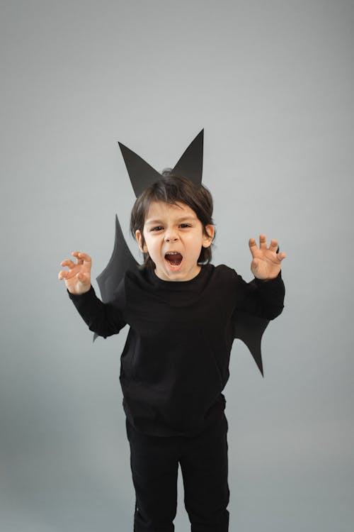 Free Angry boy in bat outfit with wings and ears looking at camera with opened mouth on gray background in studio Stock Photo