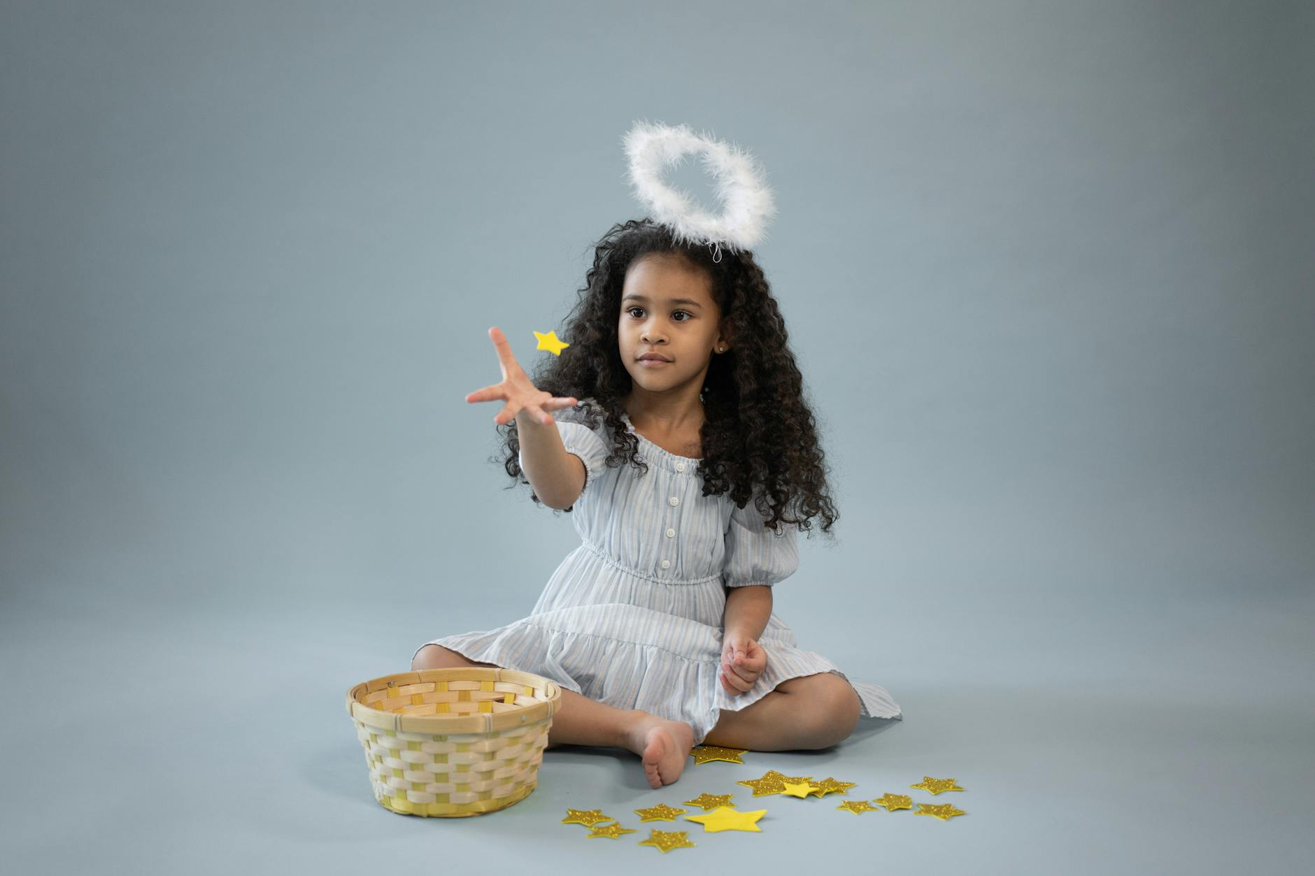 Adorable girl dressed as angel tossing stars with basket on studio floor.