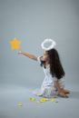 Full body of adorable African American girl in white angel outfit with halo touching decorative yellow star hanging on gray background