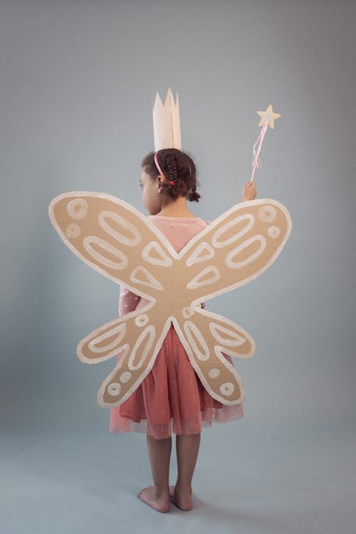 Full body back view of anonymous barefoot African American girl in fairy costume with crown and magic wand standing on gray background