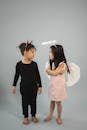 Full body of barefoot multiethnic kids wearing devil and angel outfits looking at each other on gray background in studio